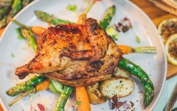 A plated dish from Spize Rifle Range featuring a roasted chicken leg quarter with crispy, seasoned skin served on a bed of roasted asparagus, carrots, and potato wedges on a light-colored plate.