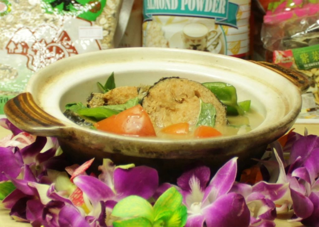 Close-up of a traditional clay pot dish containing fish, tomatoes, and green vegetables in a savory broth, garnished with fresh purple orchids.