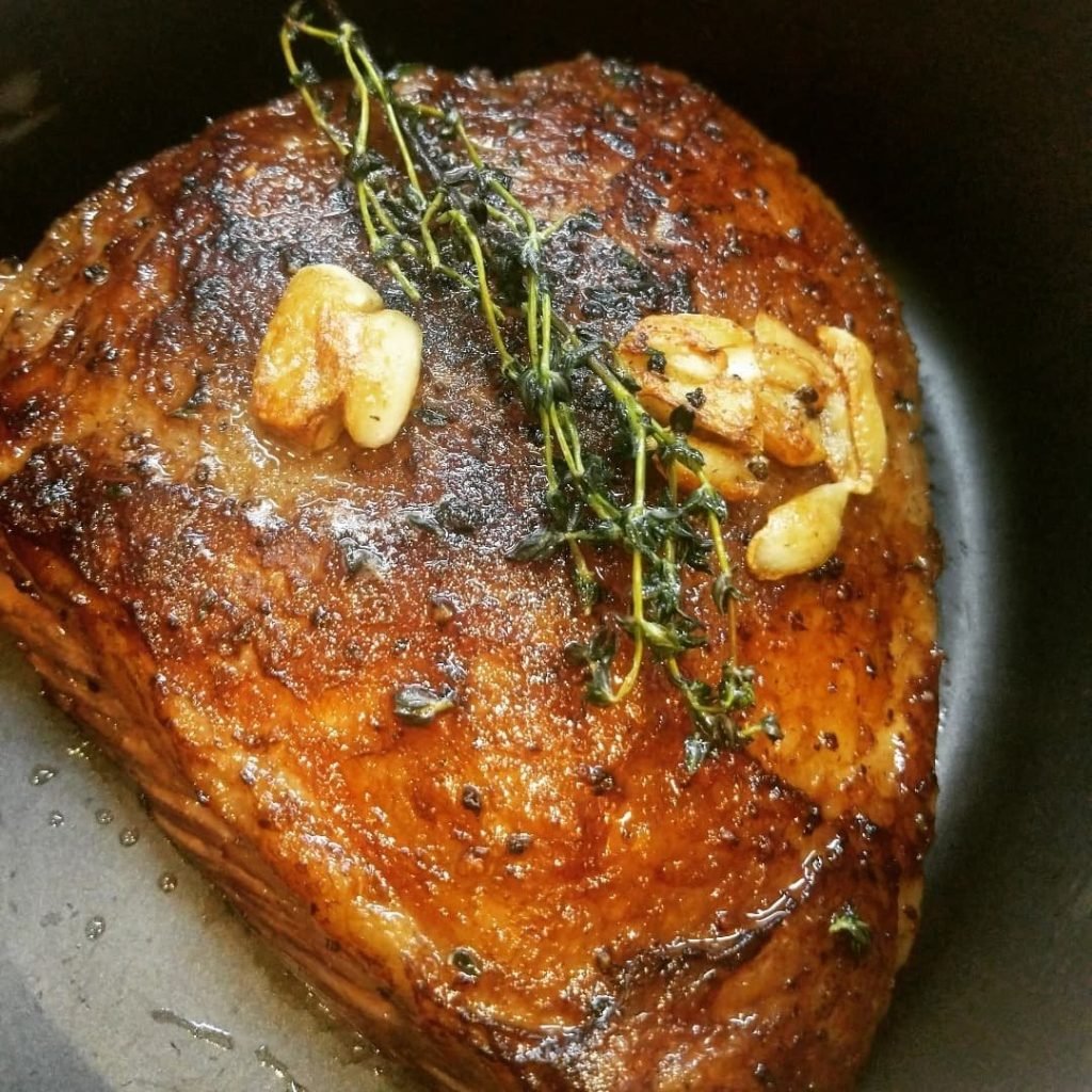 Close-up of a large beef roast, seared to a deep golden brown, topped with fresh thyme sprigs and roasted garlic cloves
