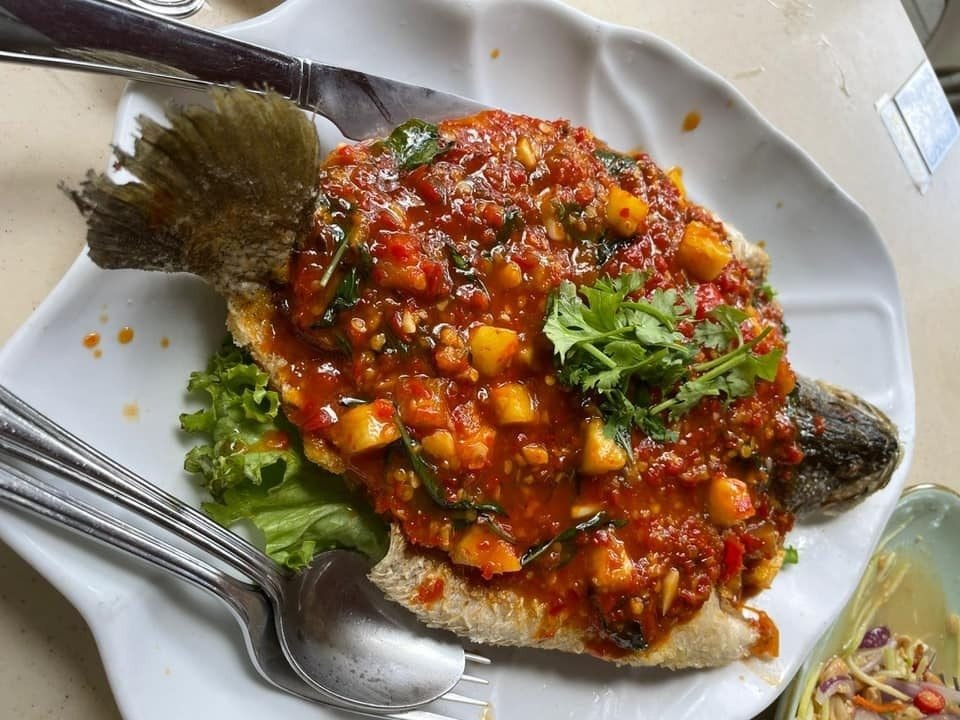 A whole deep-fried fish served on a white, wavy-edged plate, generously smothered in a vibrant red, chunky sweet chili sauce, possibly containing pineapple pieces, and garnished with fresh cilantro.