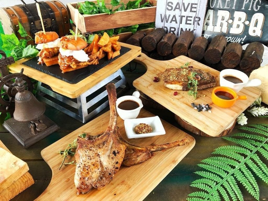 Rustic spread of grilled tomahawk pork chop, steak, sliders, and wedges, with various sauces