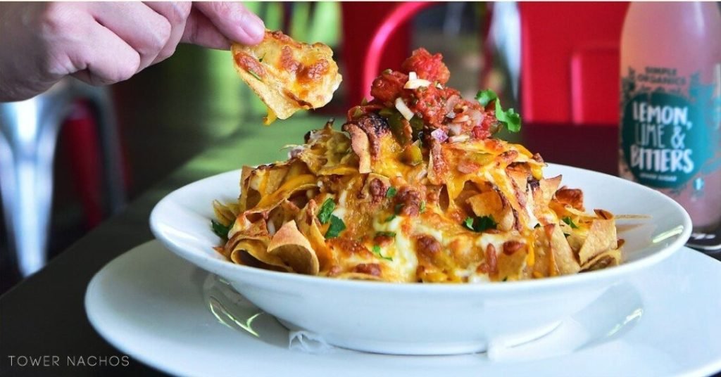 Close-up of a large white bowl piled high with 'Tower Nachos' - tortilla chips loaded with melted cheese, salsa, and cilantro, as a hand picks one up