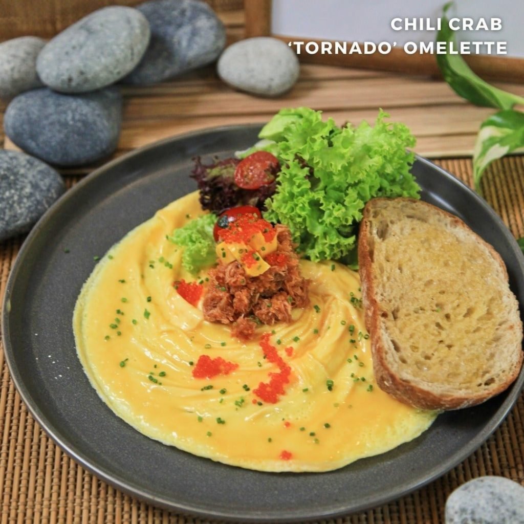 Two Bakers' 'CHILI CRAB TORNADO OMELETTE' - a swirled yellow omelette topped with chili crab meat and orange roe, served with toast and salad on a dark plate.