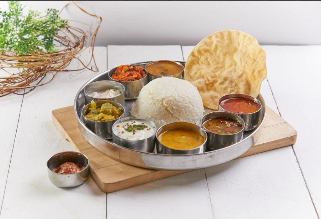 Traditional Indian Thali meal with rice, assorted curries, vegetables, yogurt, pickle, and a papadum on a steel platter.