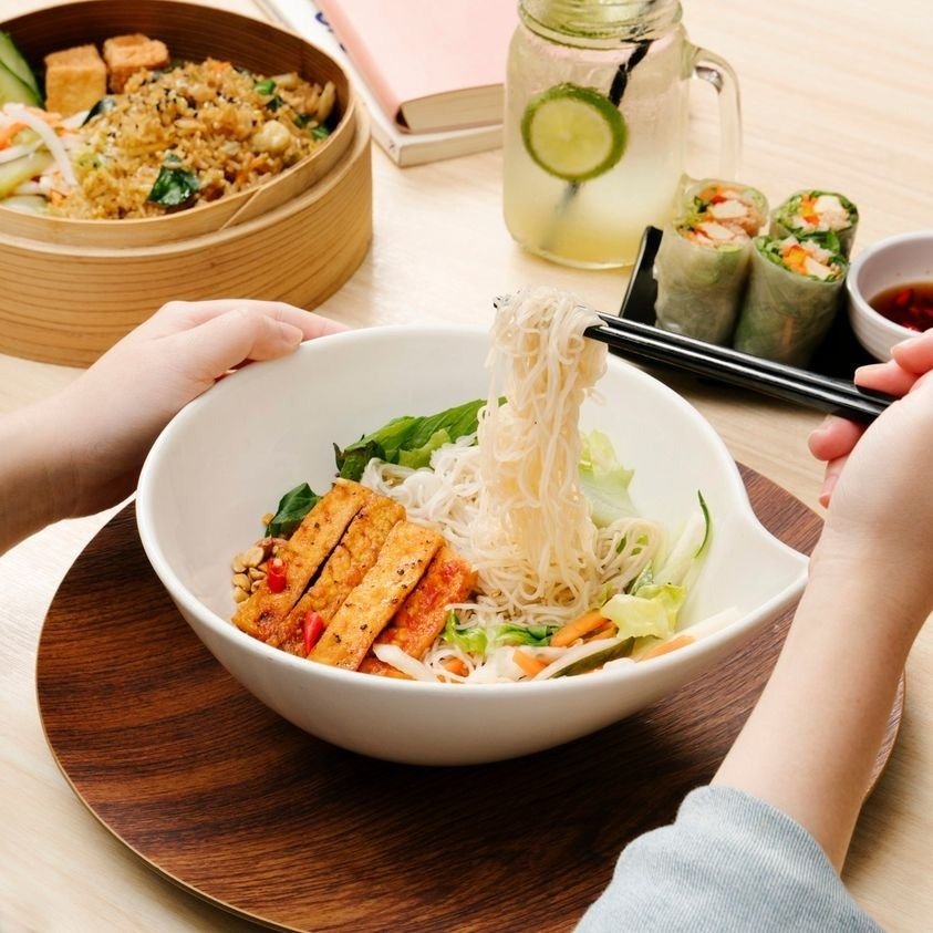 Close-up of a white bowl containing a Vietnamese-style noodle salad with tofu and vegetables, being eaten with chopsticks.