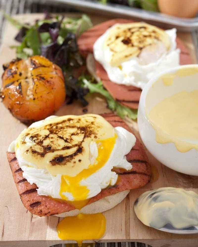 Wawawa's Eggs Benedict with grilled luncheon meat, poached eggs with runny yolk, and hollandaise sauce, served with a grilled tomato and side salad on a wooden board.