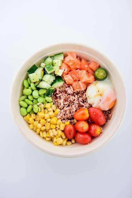 Poke bowl with salmon, rice, edamame, corn, cucumber, tomatoes, egg, and calamansi, on a white background.