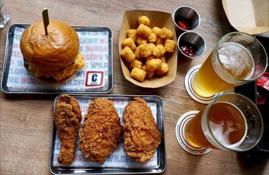 A satisfying meal at Wildfire Burgers: a juicy burger, crispy fried chicken, a side of tater tots, and two cold beers on a wooden table.