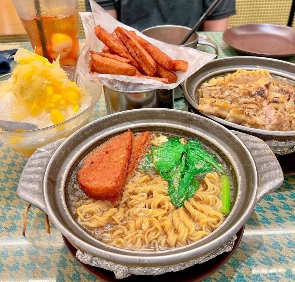 Hong Kong style meal featuring a clay pot of instant noodles with luncheon meat and vegetables, a side of grilled pork chop or chicken, luncheon meat fries, a shaved ice dessert, and an iced fruit tea.