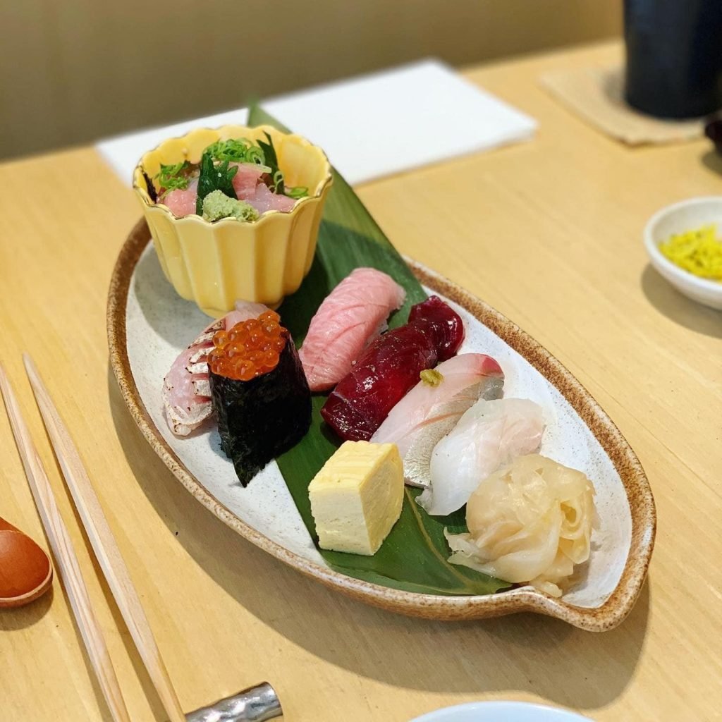 A Japanese meal set with an assortment of nigiri sushi, ikura gunkanmaki, tamagoyaki, and pickled ginger on a leaf-shaped platter, accompanied by a small bowl of chirashi don and chopsticks.