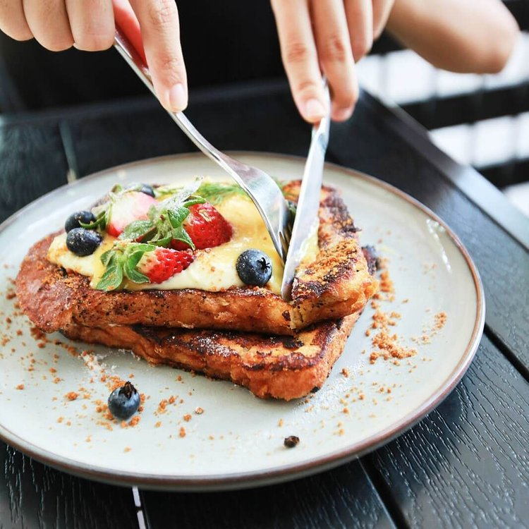 Person cutting into Daily Ground French toast topped with fresh berries, cream, and powdered sugar.