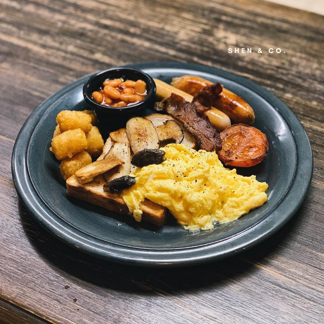 A full breakfast plate from SHEN & CO. with scrambled eggs, sausages, bacon, beans, mushrooms, tomato, and tater tots.