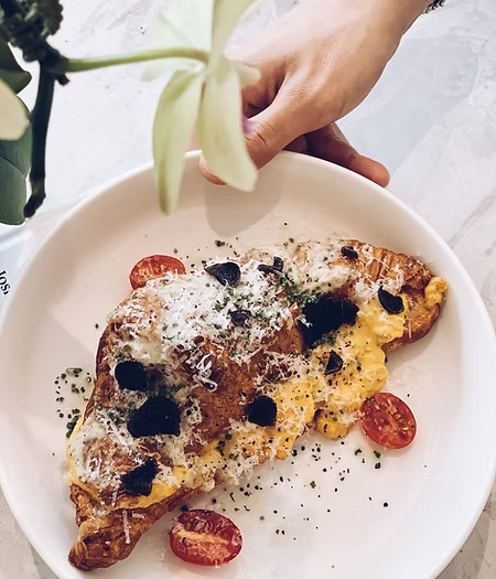 Savory croissant topped with scrambled eggs, truffle shavings, cheese, and cherry tomatoes on a white plate