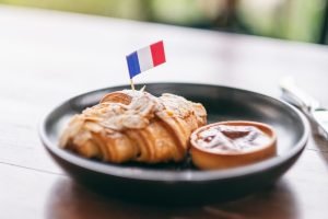An almond croissant with a small French flag on a toothpick is served on a dark plate