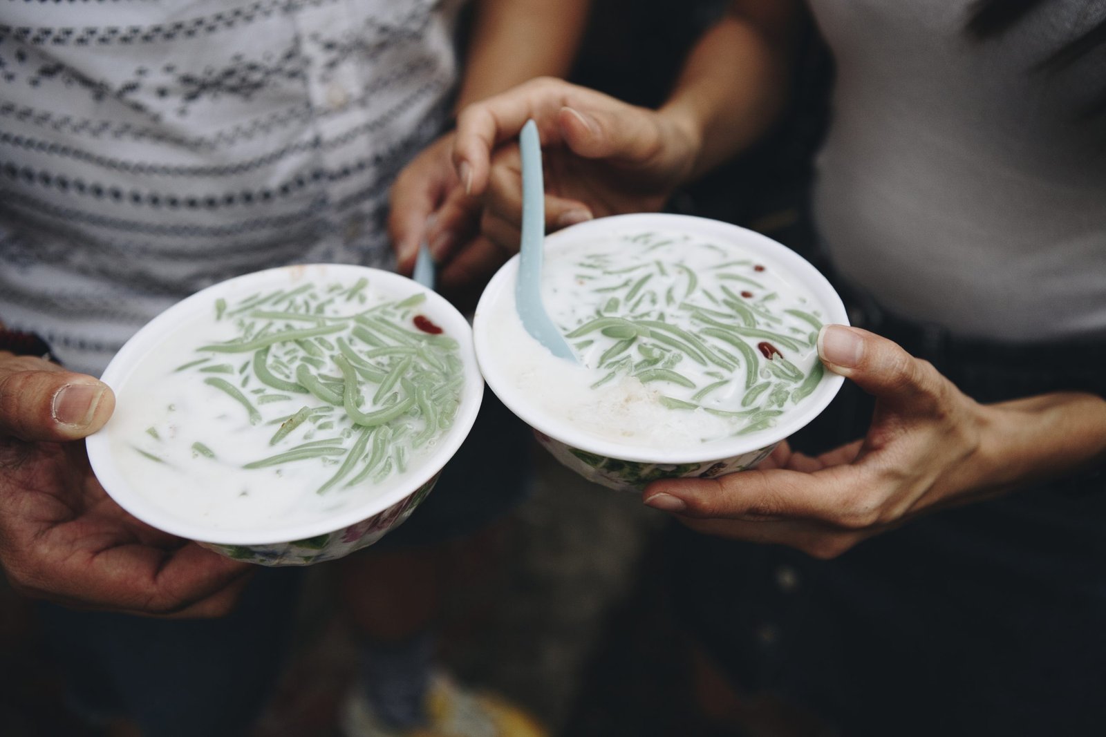 A close-up, high-angle shot of two people holding two white bowls of Cendol
