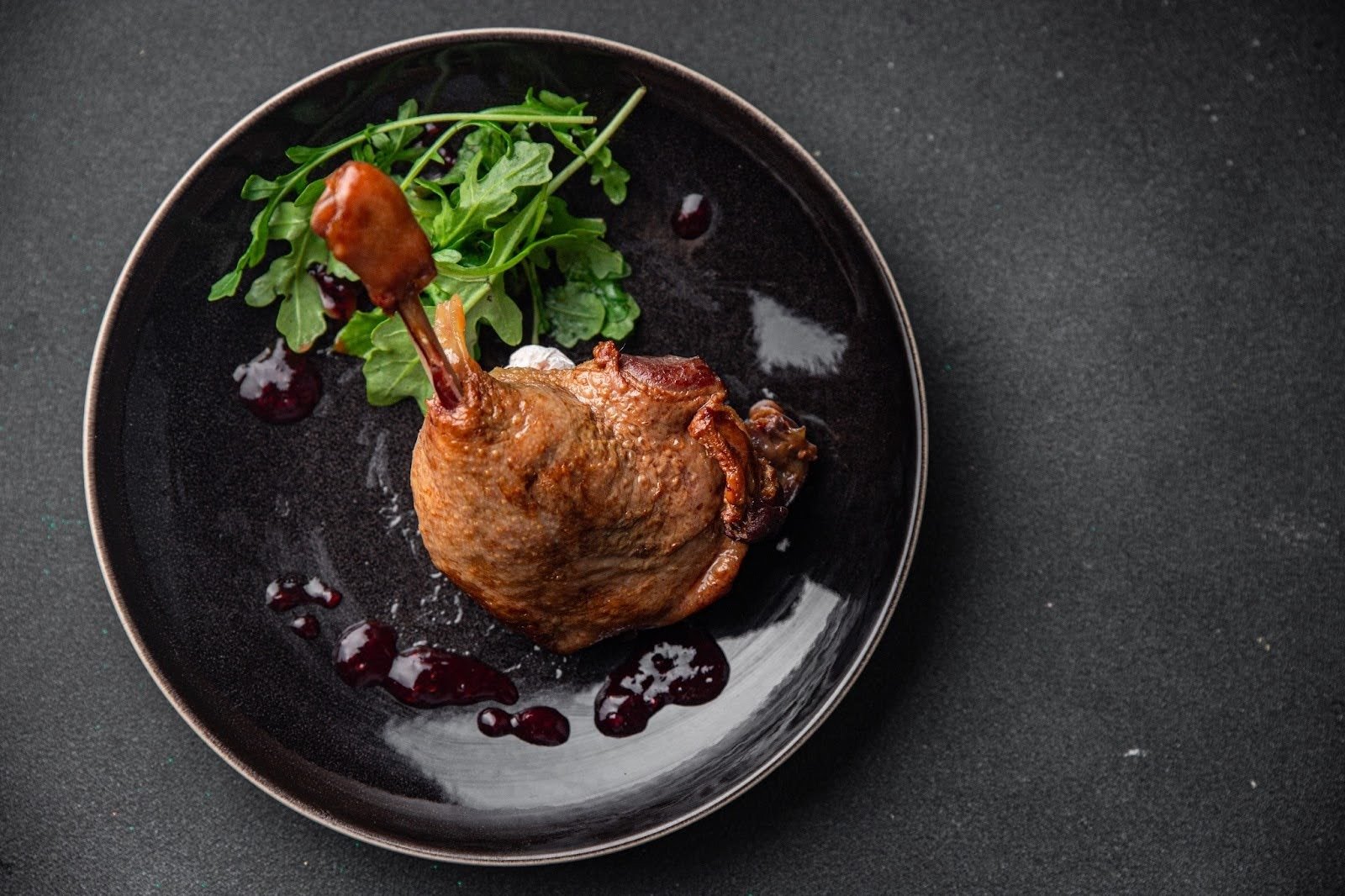 A crispy duck confit leg served on a dark plate with arugula and berry sauce
