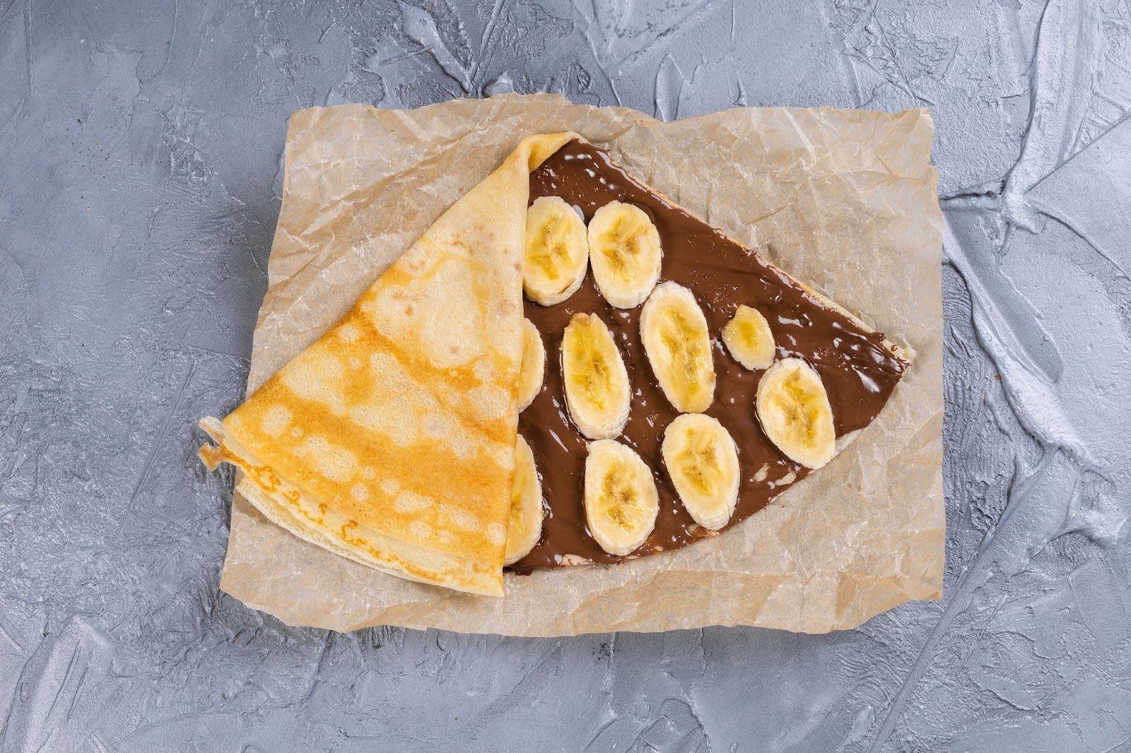 A French crêpe filled with chocolate spread and sliced bananas, being folded on parchment paper