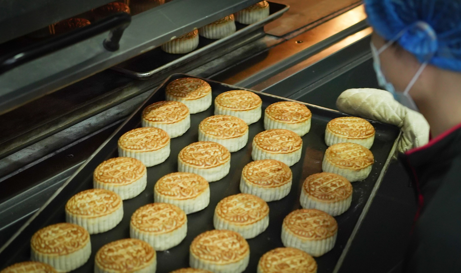 A baker at Tai Chong Kok carefully takes a tray of freshly baked, golden-brown traditional mooncakes out of a large commercial oven