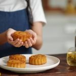 A person in a blue apron holds a traditional baked mooncake over a plate with more mooncakes and a glass of tea
