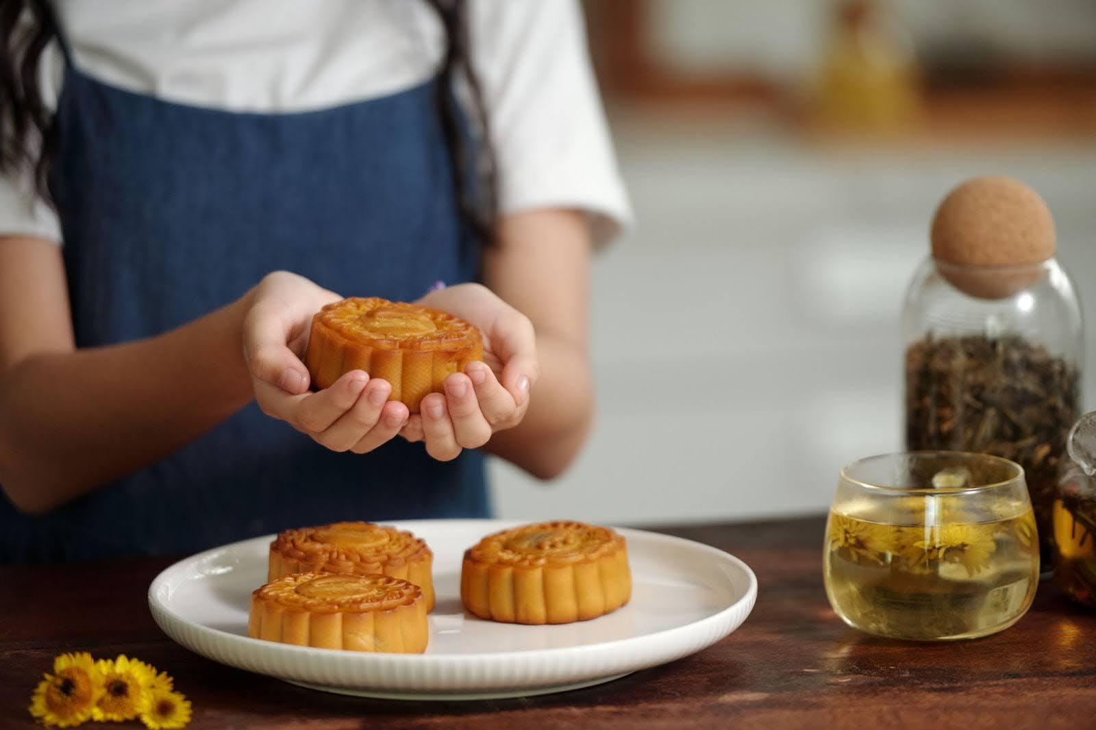 A person in a blue apron holds a traditional baked mooncake over a plate with more mooncakes and a glass of tea