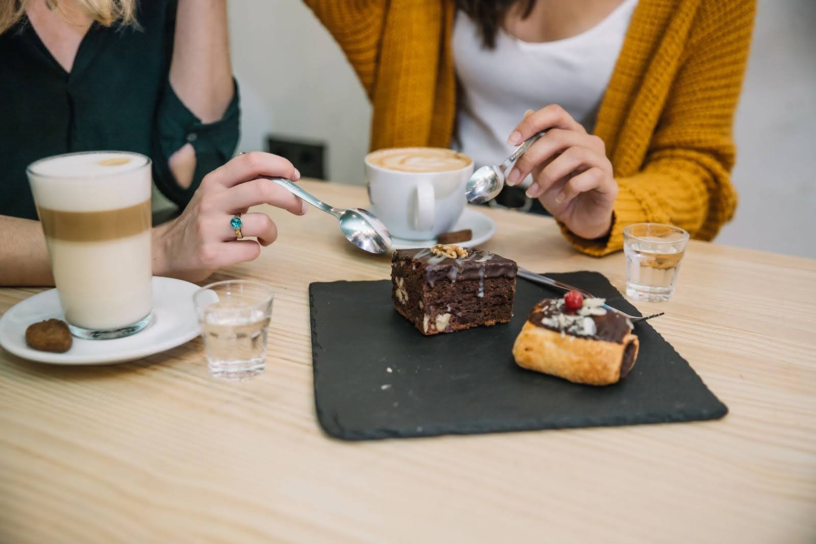 Two women sitting at a cafe table, sharing a slate platter with a brownie and a pastry while enjoying their coffee drinks