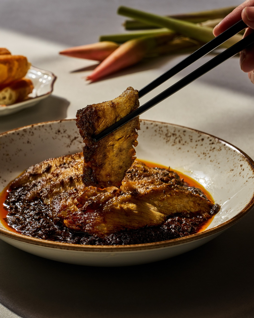 Chopsticks lifting a slice of grilled sambal stingray served in a ceramic bowl