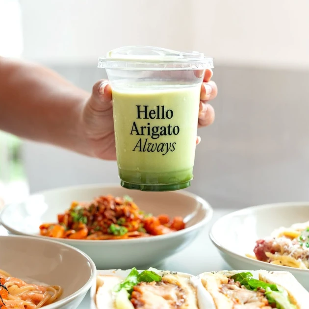 Hand holding an iced matcha latte cup above cafe table with pasta dishes and sandwiches