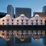 Illuminated facade of The Warehouse Hotel along Singapore River at dusk with water reflection.