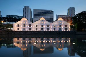 Illuminated facade of The Warehouse Hotel along Singapore River at dusk with water reflection.